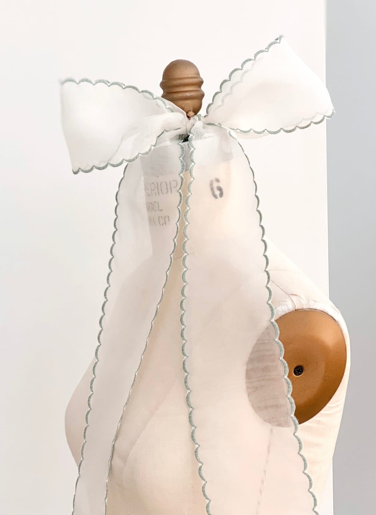 A white silk ribbon with scalloped edges is tied in a large bow atop a vintage dress form. The ribbon's delicate gray stitching contrasts with the wooden top of the mannequin, highlighting its elegance against the minimalist background.