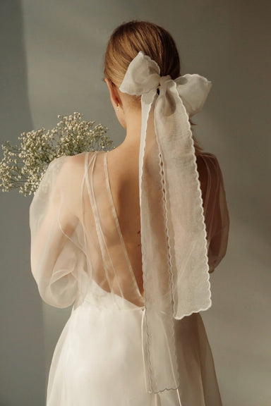 A woman with blonde hair is seen from behind, wearing a white dress with a sheer, open back. Her hair is elegantly adorned with a large bow made from Noémiah's Silk Ribbon, and she holds a bouquet of white baby's breath flowers against a neutral background.