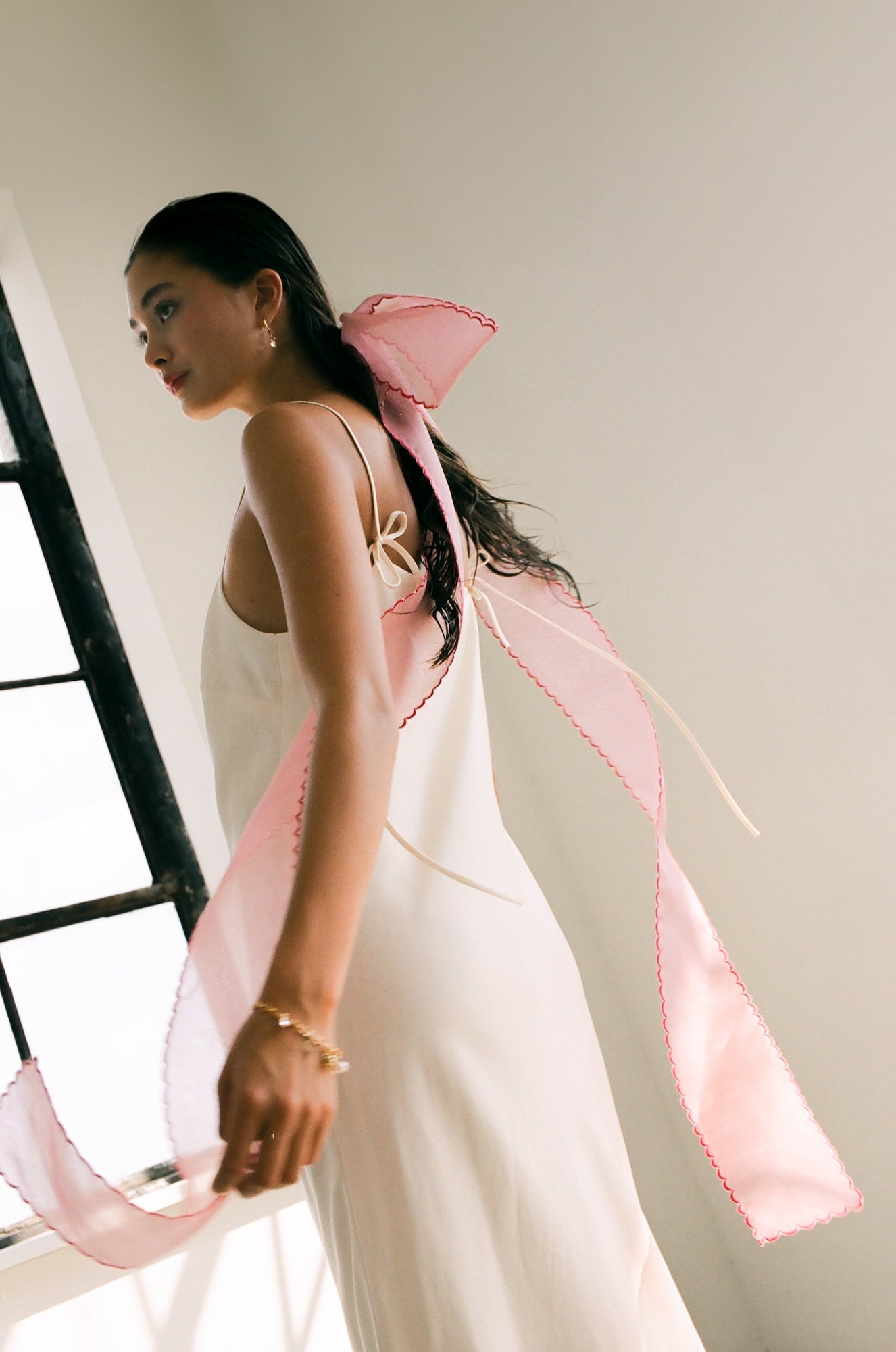 A woman, draped in a sleeveless white dress with embroidered edges, stands near a window. A large Silk Ribbon from Noémiah is tied in her hair, complementing the scene's gentle embrace of natural light.