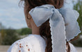 A woman with brown hair is seen from behind outdoors, wearing a white lacy open-back dress with scalloped embroidered edges and a large, light blue Noémiah Silk Ribbon — Classic in her hair against a blurred backdrop of trees and sky.