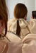 A woman with brown hair in a ponytail, styled with the Noémiah Silk Ribbon—a large cream organza bow with black ruffled edges—takes a mirror selfie while wearing a matching sheer top.