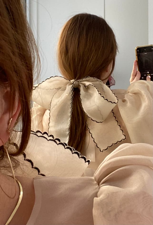 A woman with brown hair in a ponytail, styled with the Noémiah Silk Ribbon—a large cream organza bow with black ruffled edges—takes a mirror selfie while wearing a matching sheer top.