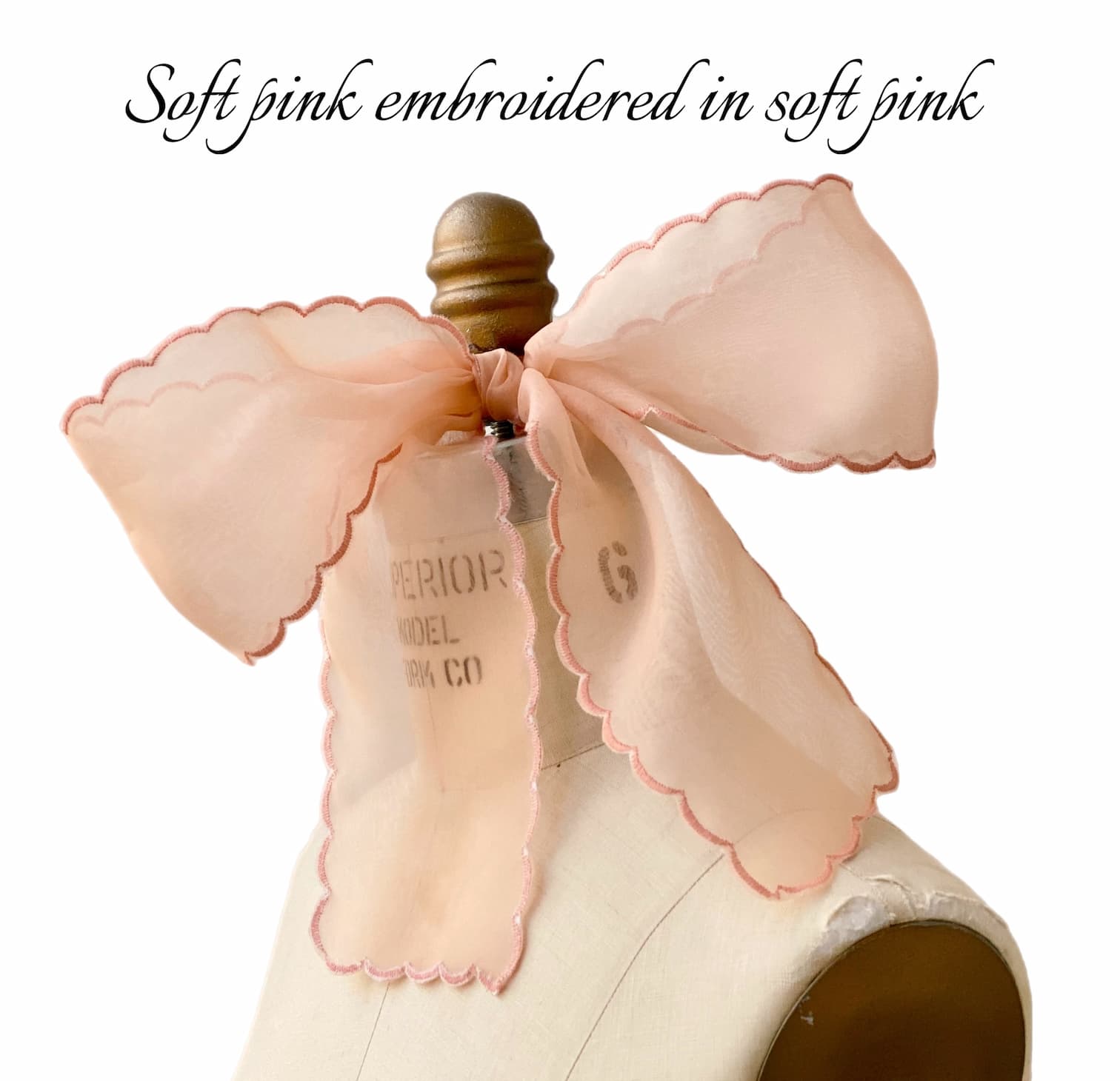 A soft pink Silk Ribbon by Noémiah with scalloped edges is tied around a dress form’s neck. The sheer organza fabric features matching pink embroidery. Upper text: “Soft pink embroidered in soft pink.”.