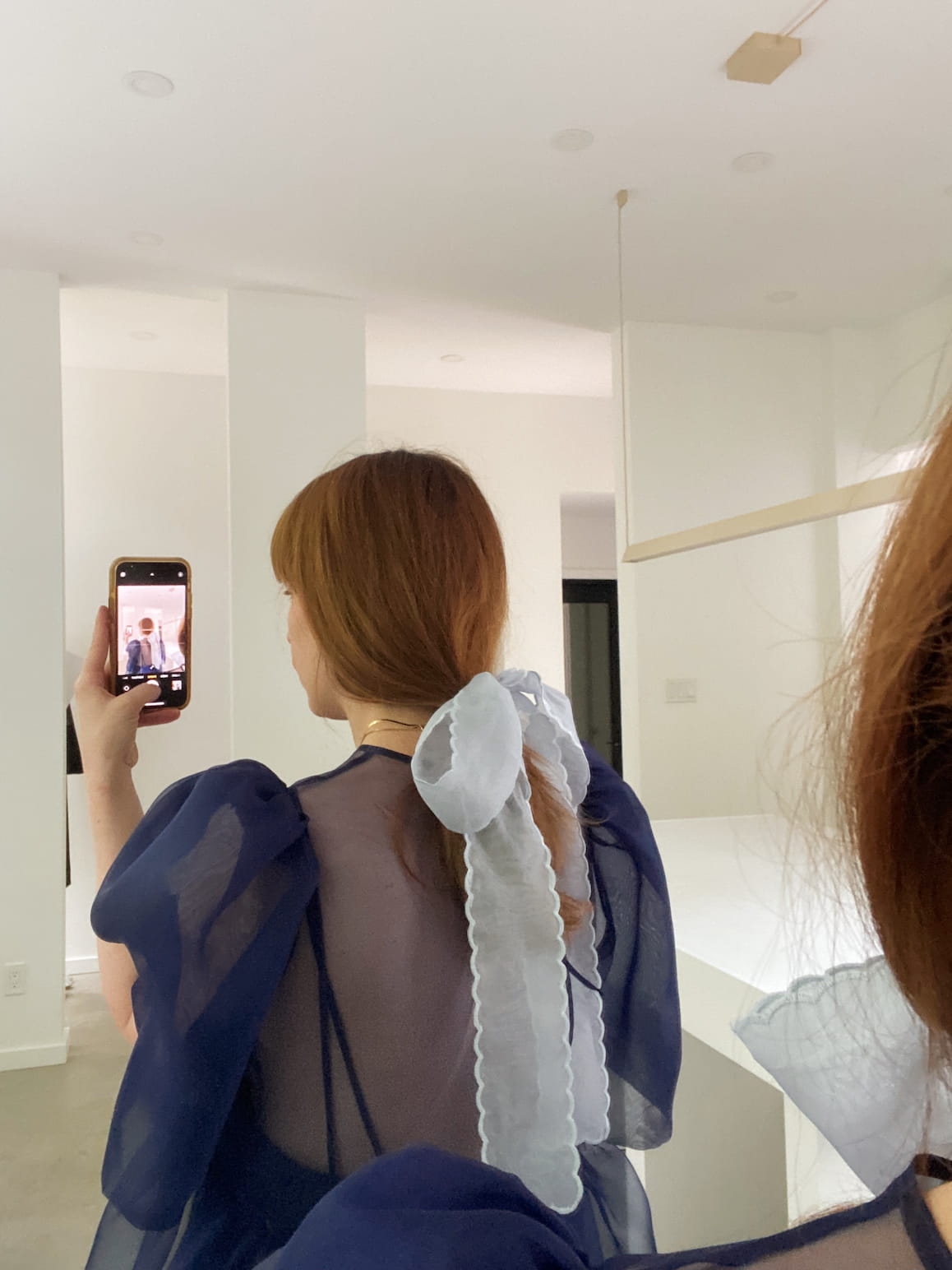 A woman with long brown hair, tied back with the Noémiah Silk Ribbon — Classic, takes a mirror selfie in a bright modern room while wearing a sheer dark blue puffy-sleeved top.