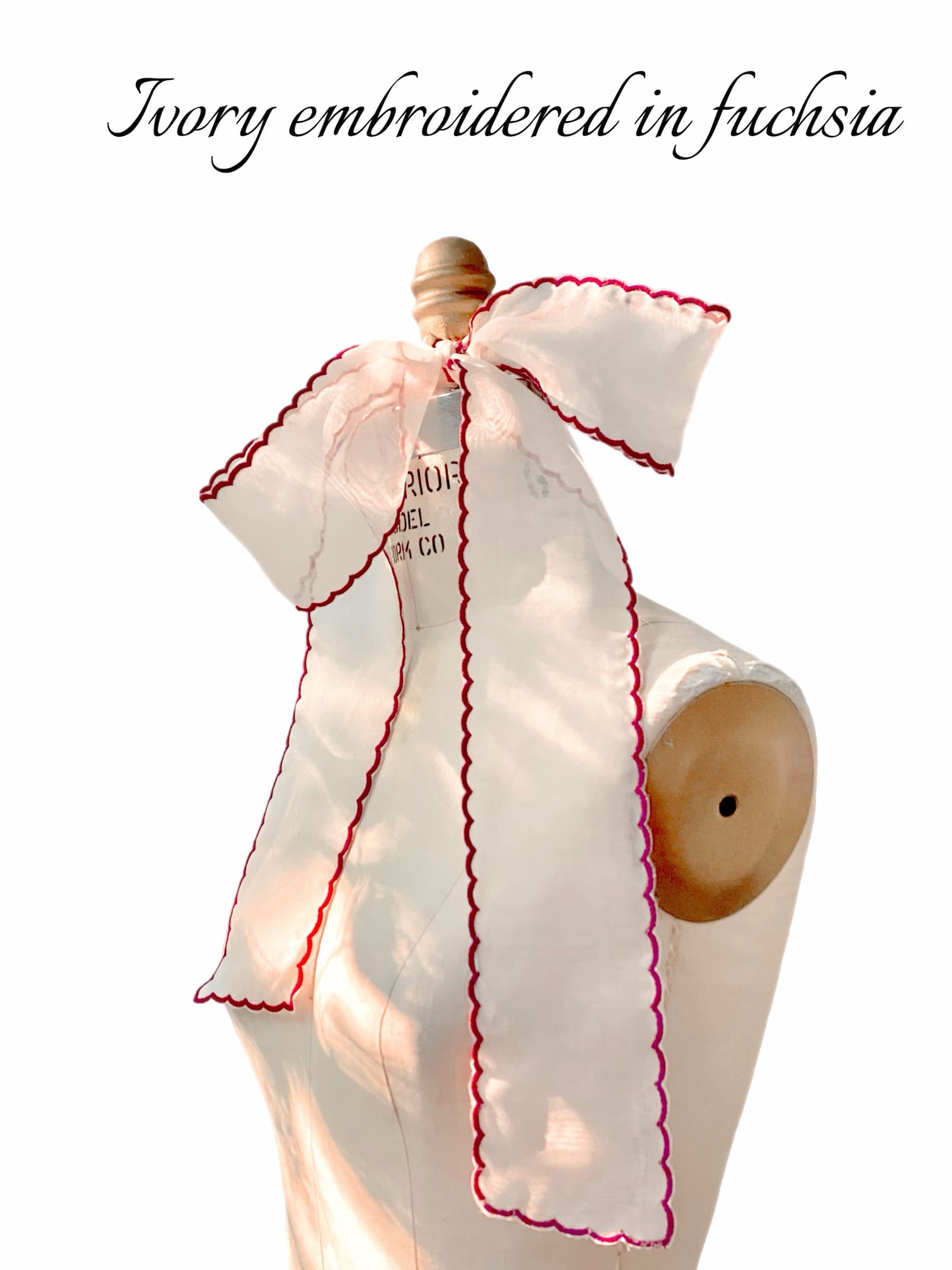 Noémiah’s Silk Ribbon in ivory organza features fuchsia scalloped embroidery and is tied in a large bow, as shown on a dress form. The text above reads, “Ivory embroidered in fuchsia.” An elegant accessory for any look.