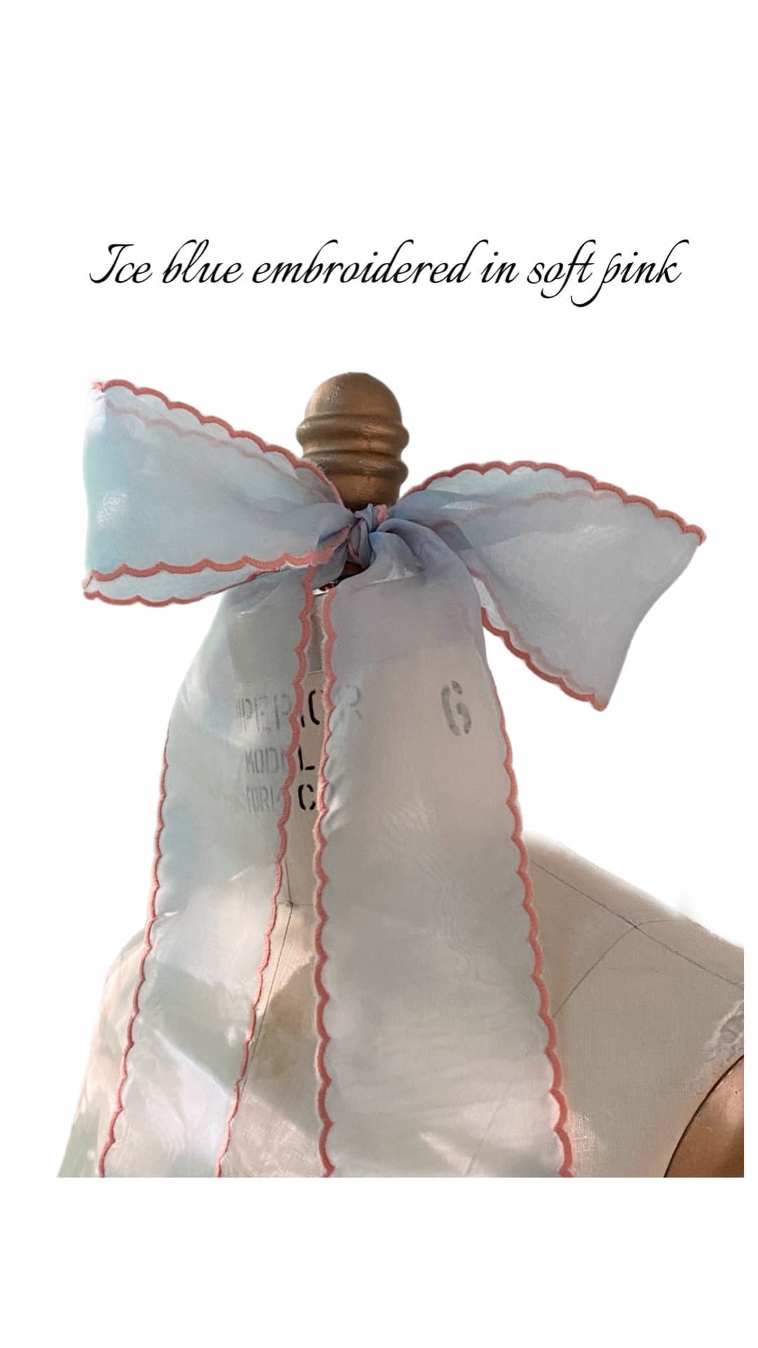 A large, sheer Noémiah Silk Ribbon in ice blue with soft pink scalloped embroidery is tied in a bow around the top of a wooden dress form. Text above reads: Ice blue embroidered in soft pink.