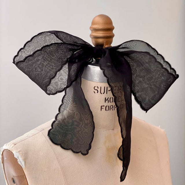 A mannequin bust is adorned with the Silk Ribbon — Classic from Noémiah. The large, sheer black bow tied around the neck features scalloped embroidered edges and delicate lace-like patterns, adding a touch of elegance against the blurred indoor background.