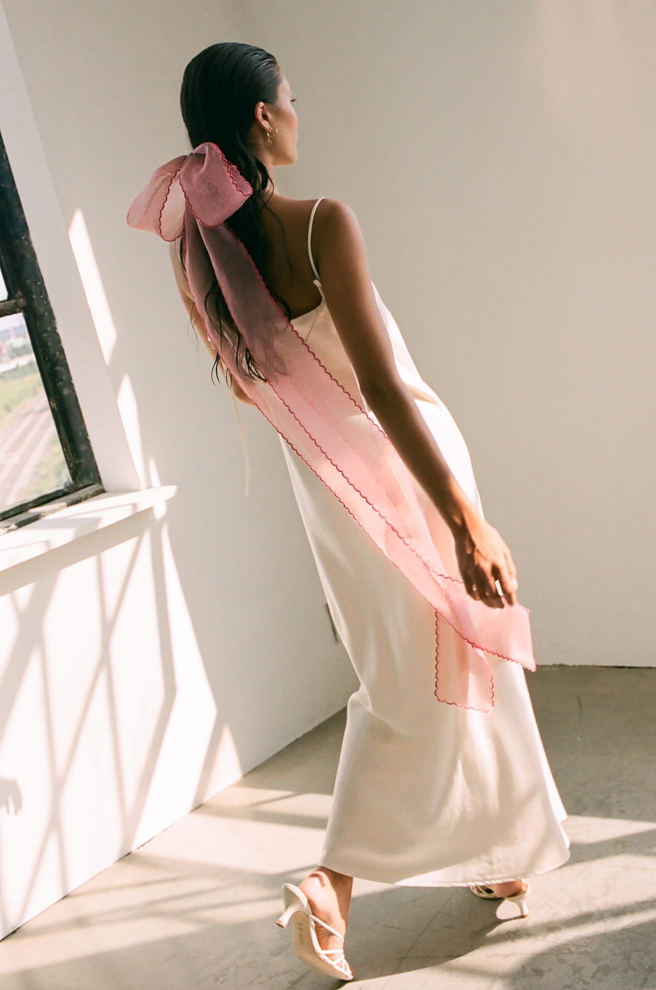 A woman in a long white slip dress and heeled sandals stands by a sunlit window. Her hair is adorned with the Noémiah Silk Ribbon — Classic, the pink organza trailing gracefully down her back as soft natural light fills the space.