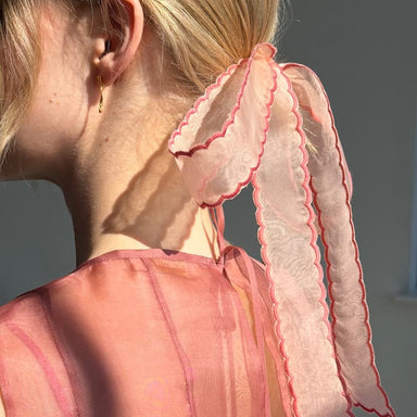 A woman with blonde hair in a low ponytail is adorned by the Noémiah Silk Ribbon — Thin, a sheer pink ribbon with scalloped edges and red trim. She wears a sheer pink top and small gold hoop earrings as sunlight highlights her look.