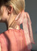 A woman with blonde hair in a low ponytail is adorned by the Noémiah Silk Ribbon — Thin, a sheer pink ribbon with scalloped edges and red trim. She wears a sheer pink top and small gold hoop earrings as sunlight highlights her look.