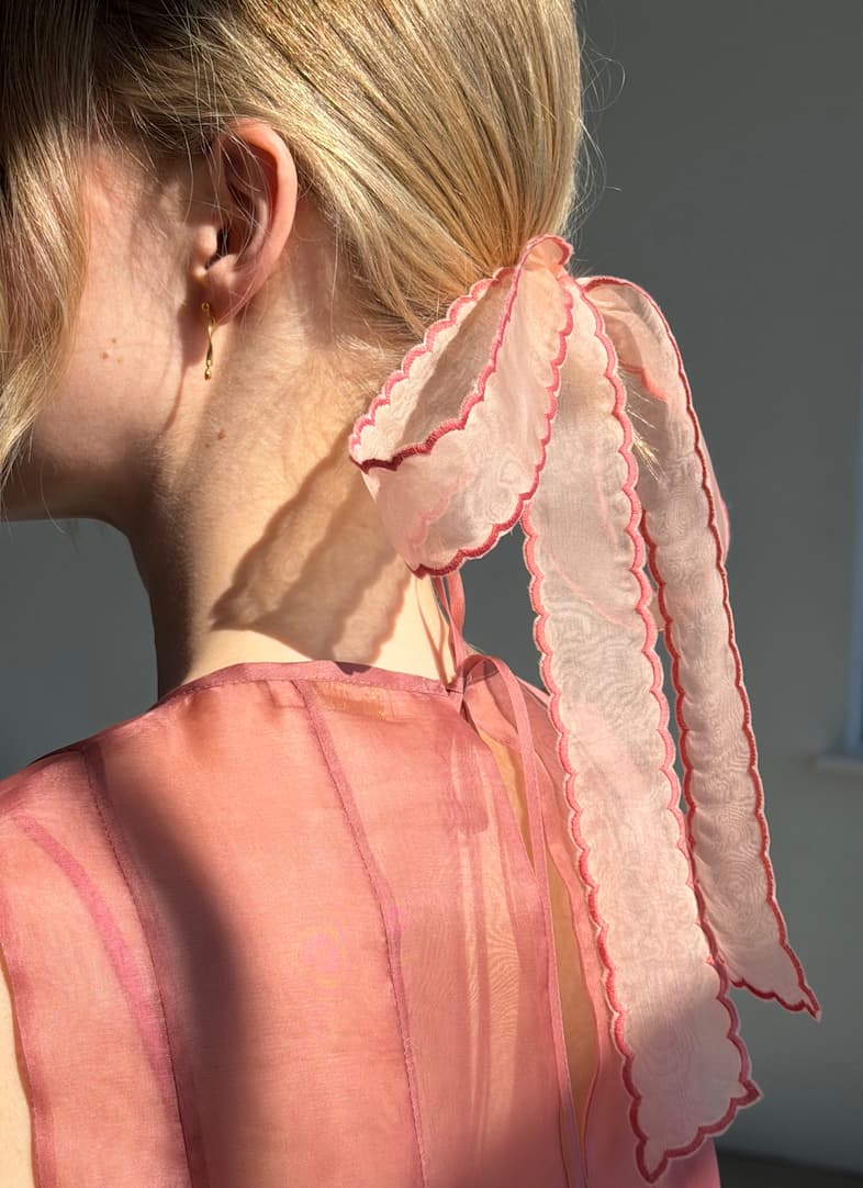 A woman with blonde hair in a low ponytail is adorned by the Noémiah Silk Ribbon — Thin, a sheer pink ribbon with scalloped edges and red trim. She wears a sheer pink top and small gold hoop earrings as sunlight highlights her look.