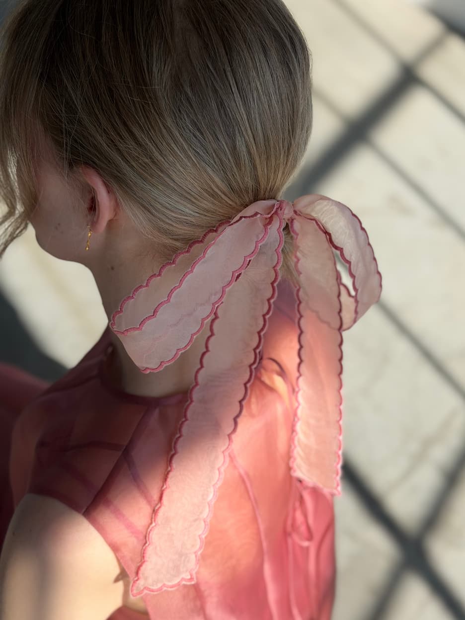A woman with light brown hair is seen from behind, wearing a sheer pink top and a delicate Noémiah Silk Ribbon — Thin tied in her hair. Sunlight casts patterned shadows on the floor in the background.