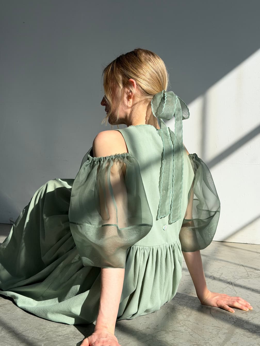 A woman with blonde hair tied in a Noémiah Silk Ribbon — Thin sits on the floor in sunlight, wearing a sage green dress with puffed, sheer sleeves and open shoulders, facing away from the camera in a minimalist space.