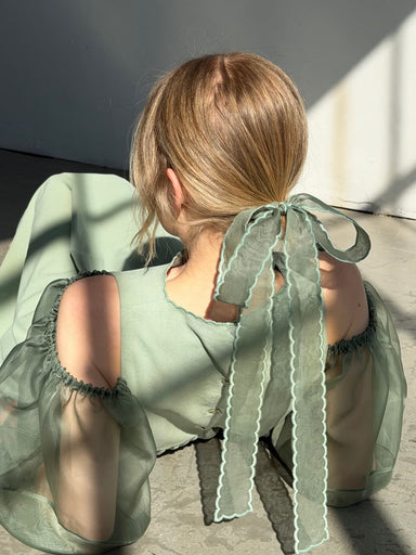 A person with blonde hair, wearing a light green dress with sheer puffed sleeves and scalloped edges, has a Noémiah Silk Ribbon — Thin tied in their hair. Sunlight creates shadows as they sit on the floor, facing away.