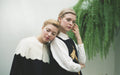 Two people stand close together, one resting their head on the other's shoulder. Both have glittery eye makeup and wear formal clothing, with one of them adorned in the elegantly embroidered Simone Top by Noémiah. A lush green fern serves as the backdrop for this intimate moment.