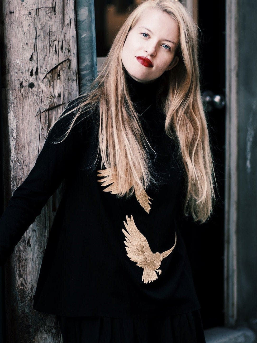 A person with long blonde hair stands against a wooden post, wearing the Simone Top by Noémiah, which is embellished with elegant gold bird designs. The background is slightly blurred to keep the focus on the person.