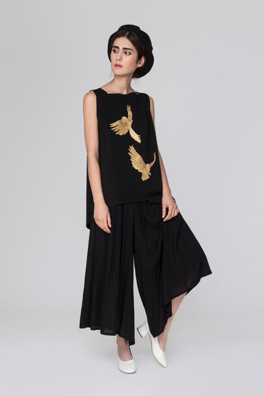 A woman stands against a gray background, wearing a Noémiah Simone top featuring gold bird designs on the embroidered fabric. She pairs it with wide black A-line pants and a black beret. Her white shoes complete the look as she gazes to the side with a relaxed pose.