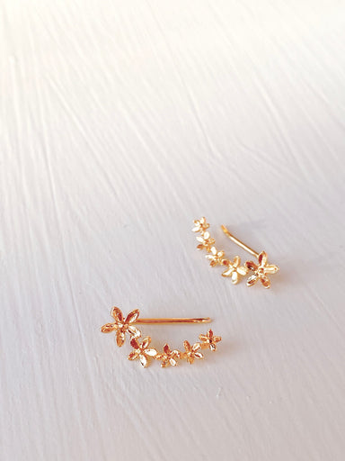 The Sisi Earrings by Noémiah, featuring small flower motifs arranged in a curved design, are showcased on a white textured surface. These tarnish-resistant accessories boast a delicate floral pattern that adds an elegant touch.