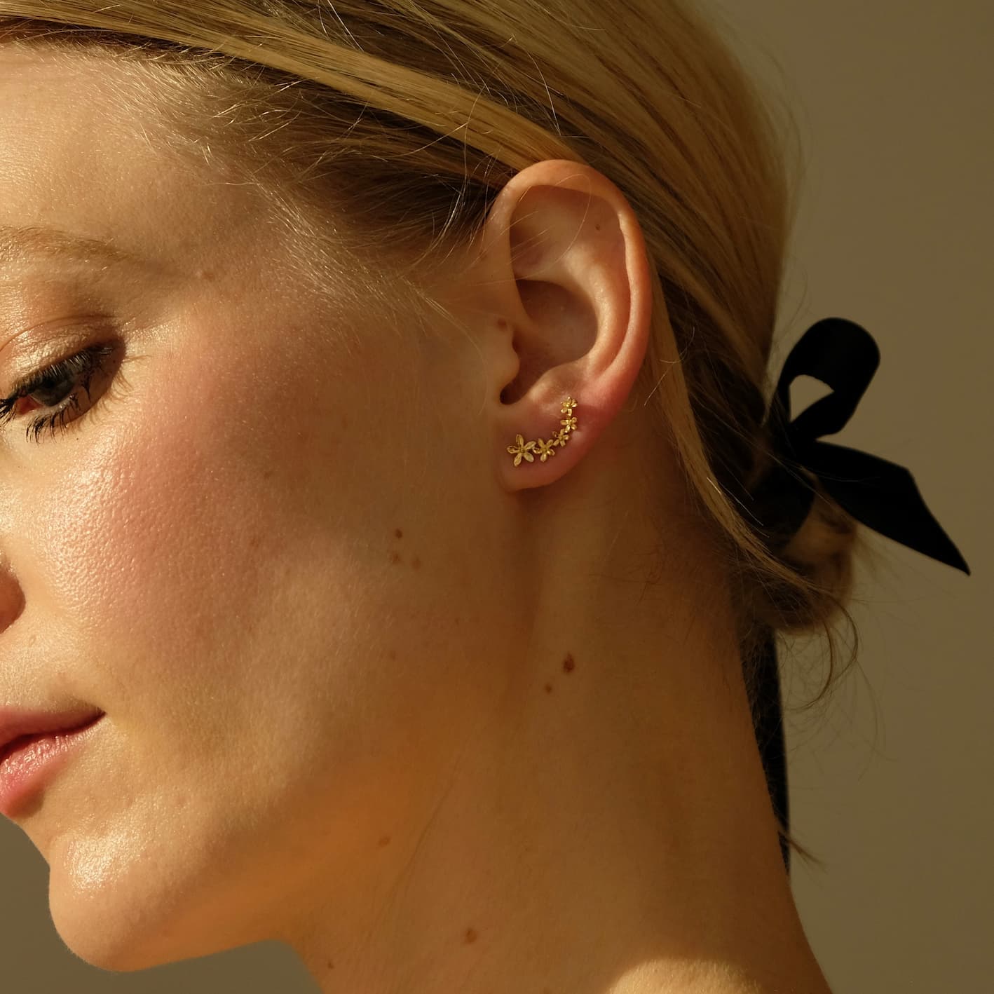 A woman with blond hair tied in a black ribbon gazes downward, her ears adorned with the delicate Sisi Earrings by Noémiah. The tarnish-resistant gold floral design is highlighted by the warm lighting, showcasing their exquisite detail against her skin.