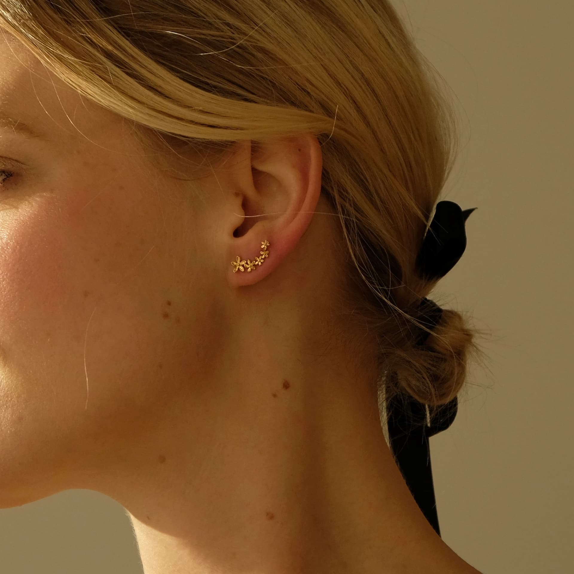 A woman with blonde hair tied in a low bun adorned with a black ribbon is wearing tarnish-resistant gold Sisi Earrings by Noémiah, featuring dainty floral designs on her right ear. The image is softly lit, creating a warm and elegant atmosphere.