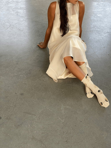 A person sits on the floor in a sleeveless white dress made of organic cotton, paired with Noémiah's Socks — Flowers Hand Dyed Pattern in white with black spots. Their long hair drapes down as they lean back, supported by one arm, with only their torso and legs visible.