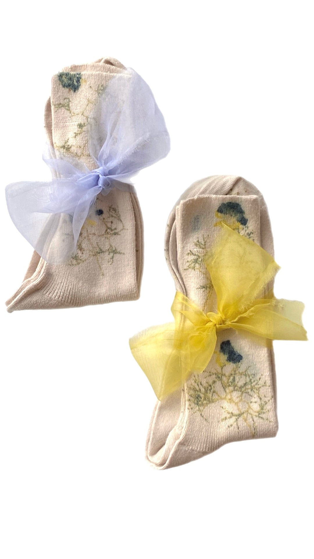 The limited edition set of Noémiah's "Socks — Flowers Hand Dyed Pattern" includes two pairs of beige socks, each featuring a delicate floral design crafted from organic cotton. One pair is tied with a soft, translucent purple ribbon, and the other with a yellow ribbon, all presented against a plain white background.