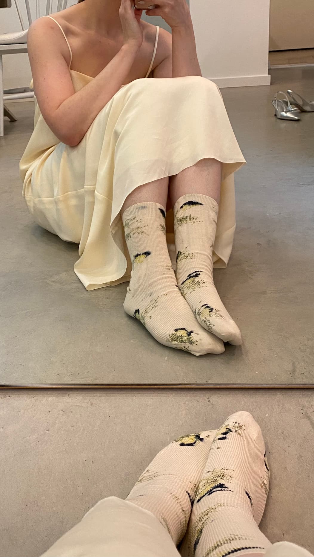 A person in an off-white dress sits on the floor, capturing a mirror selfie. They are wearing Noémiah Socks with a floral hand-dyed pattern in light beige made from organic cotton. In the background, there's a hint of style with limited edition silver high-heeled shoes visible.