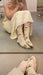 A person in an off-white dress sits on the floor, capturing a mirror selfie. They are wearing Noémiah Socks with a floral hand-dyed pattern in light beige made from organic cotton. In the background, there's a hint of style with limited edition silver high-heeled shoes visible.