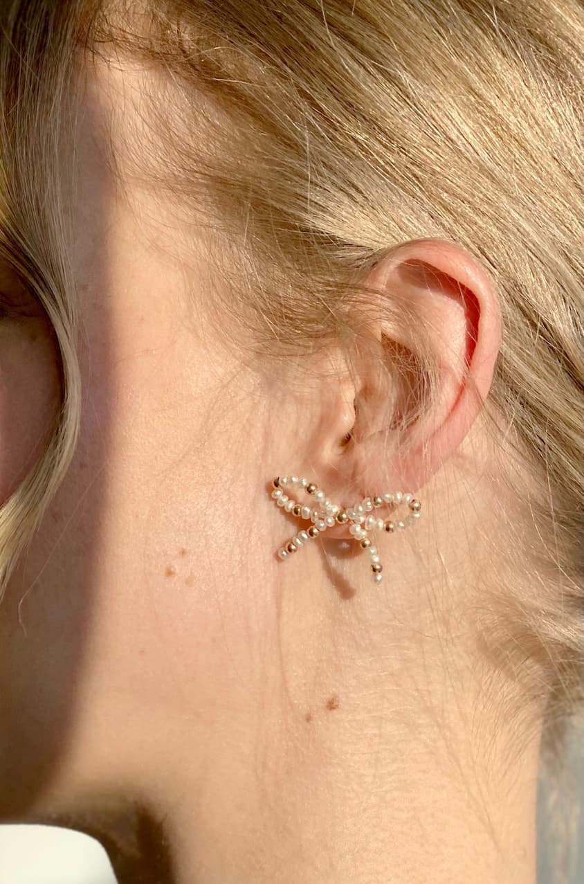 A close-up of a person's ear showcases the exquisite Sol Earrings, designed by Noémiah. These decorative earrings are shaped like branches, crafted from 14K solid gold, and adorned with small freshwater pearls. The blurred background enhances the focus on the earring and the person's light brown hair, highlighting Noémiah's remarkable craftsmanship.