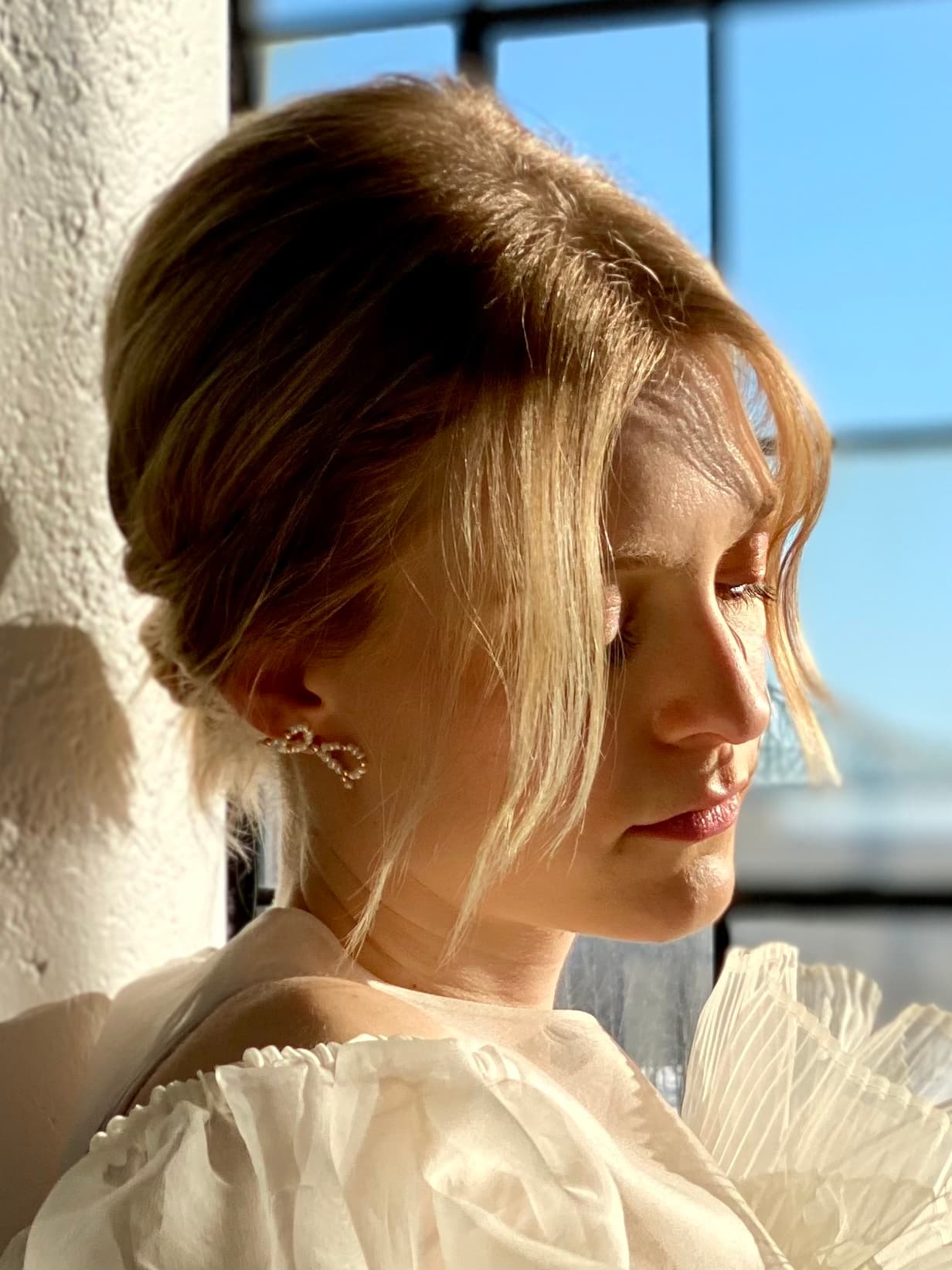 A woman with blonde hair styled in a loose updo faces right, her eyes closed and gently illuminated by soft sunlight. She is elegantly dressed in a white gown featuring freshwater pearls near the shoulders. The scene is set against a large window showcasing a clear blue sky, reflecting the refined aesthetic of Noémiah's Sol Earrings.