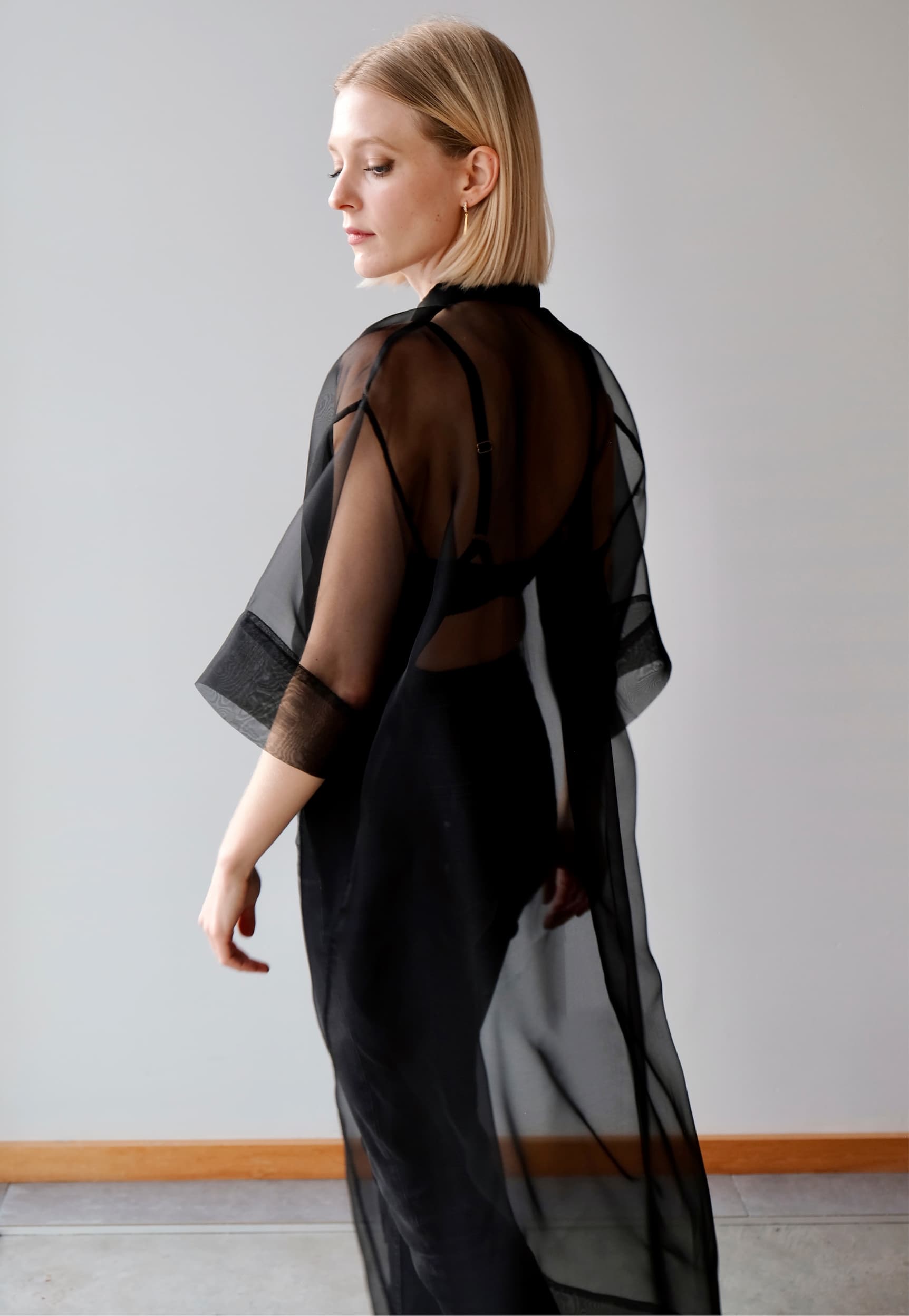 A person with blonde hair stands sideways, draped in the sheer elegance of the Sophia Classic Shirt Dress by Noémiah, featuring a black silk organza design over darker undergarments that embraces an oversized fit silhouette against a plain light gray background.