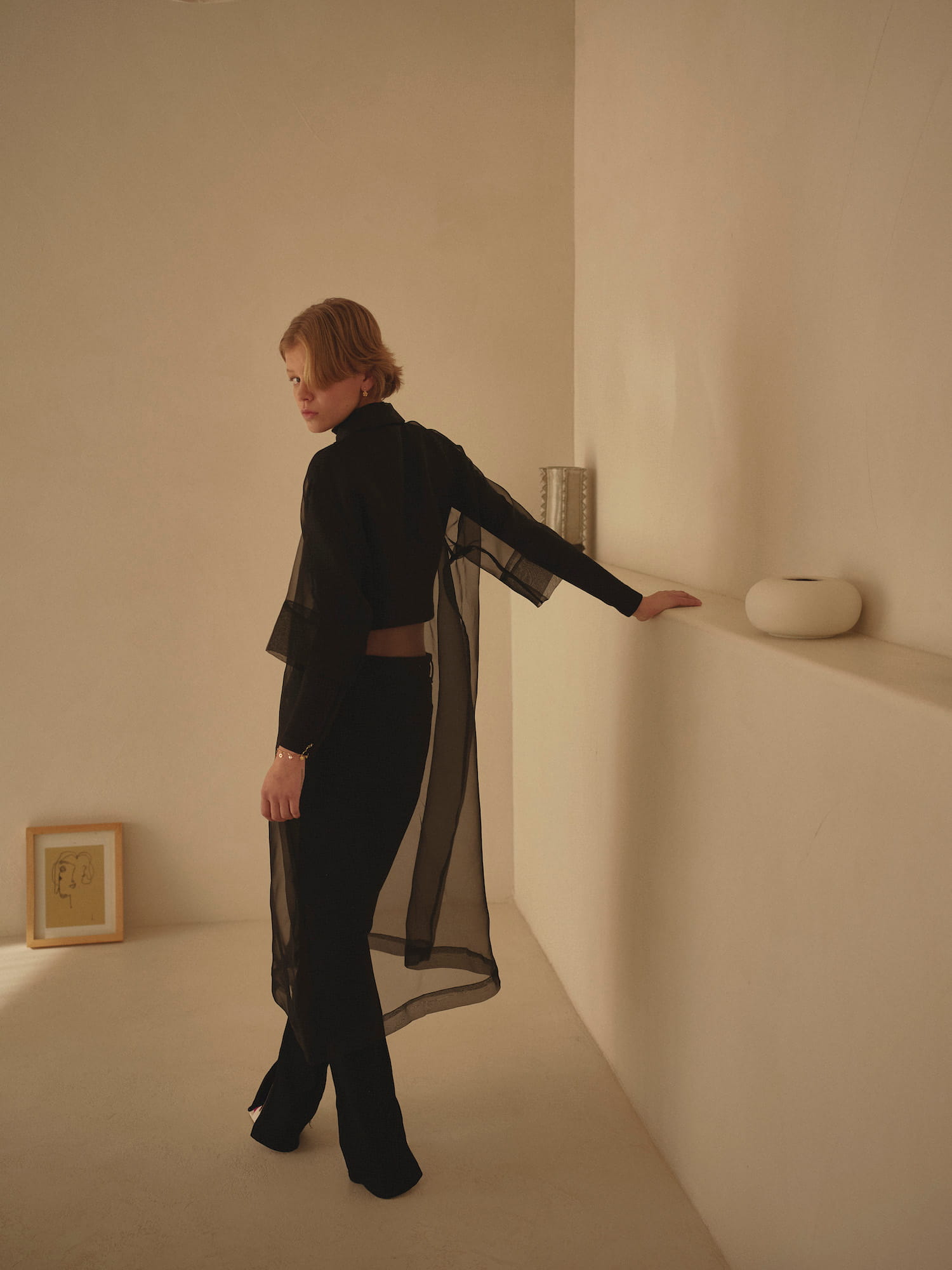 A person in a sheer black Sophia Classic Shirt Dress by Noémiah stands in a minimalist room with neutral tones. The oversized fit complements the serene setting. A white vase and framed art on the shelf add a touch of decor as they glance back over their shoulder.