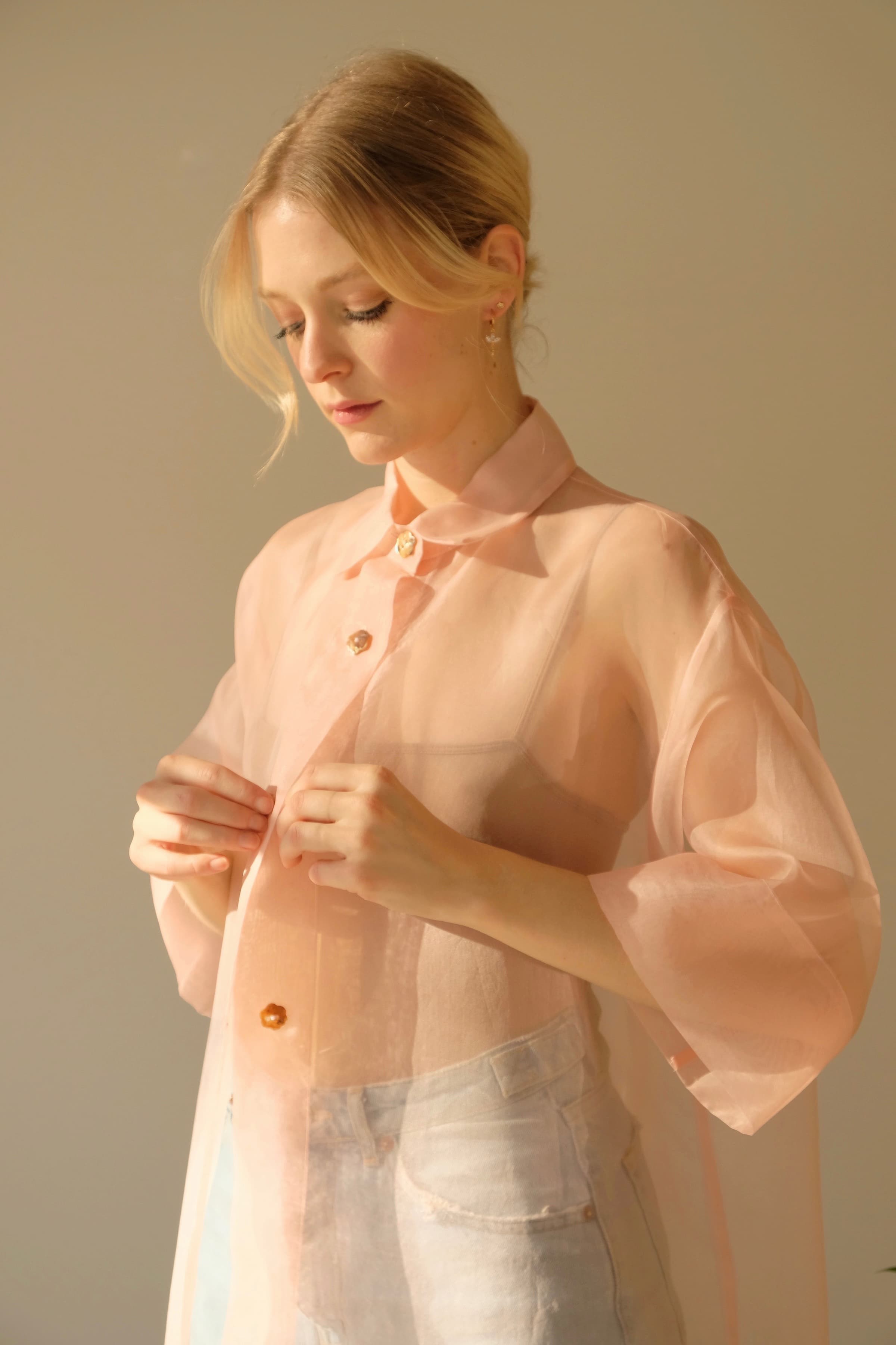 A woman with blonde hair is wearing a sheer, silk organza blouse featuring voluminous sleeves and button details. She adjusts the buttons of her Noémiah Sophia Classic Shirt Dress while standing against a soft, neutral background.