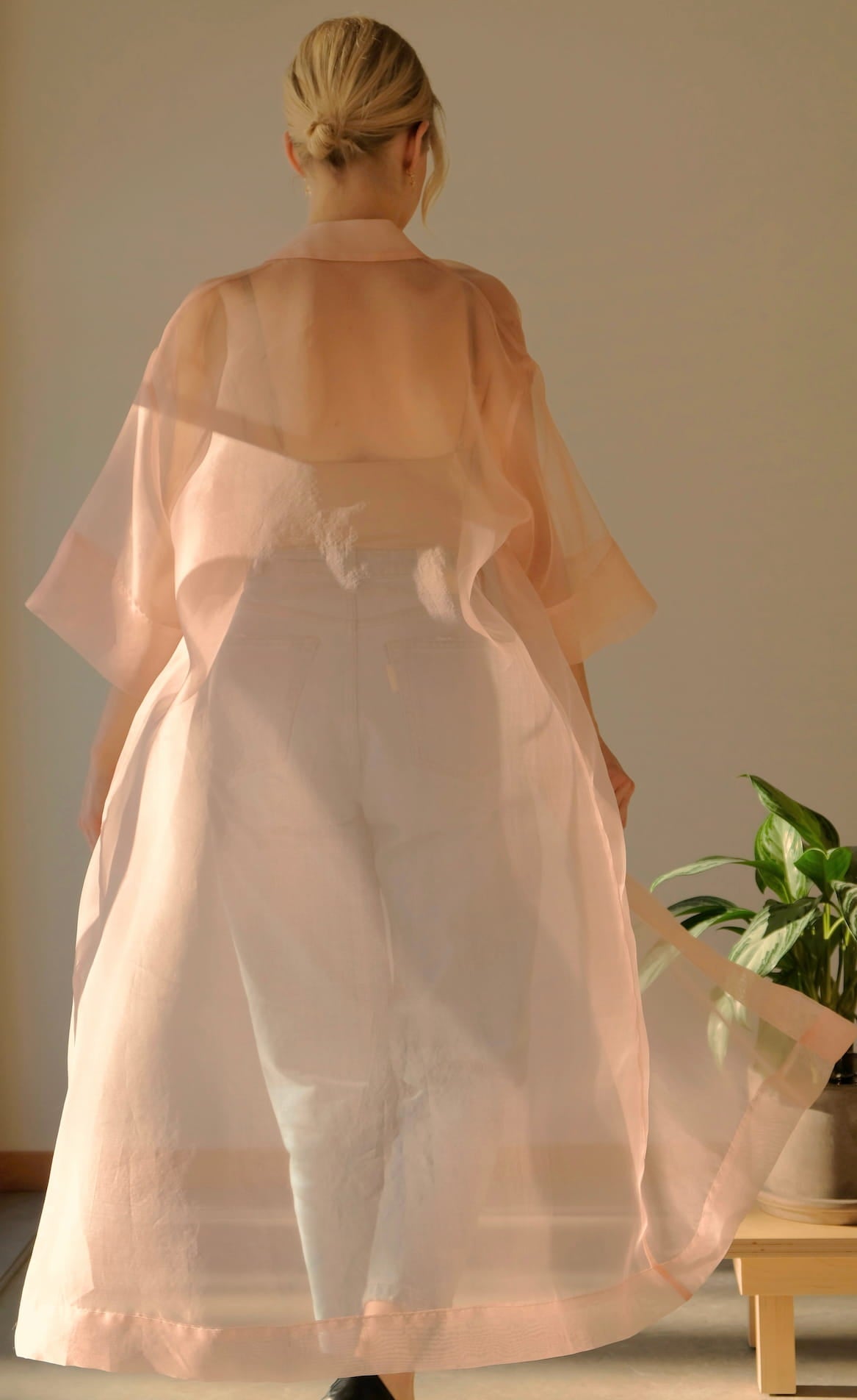 With their back to the camera, a person with blonde hair dons the Noémiah Sophia Classic Shirt Dress, wearing it over white oversized-fit pants. Natural light softly bathes the scene while a small potted plant rests on the nearby wooden floor.