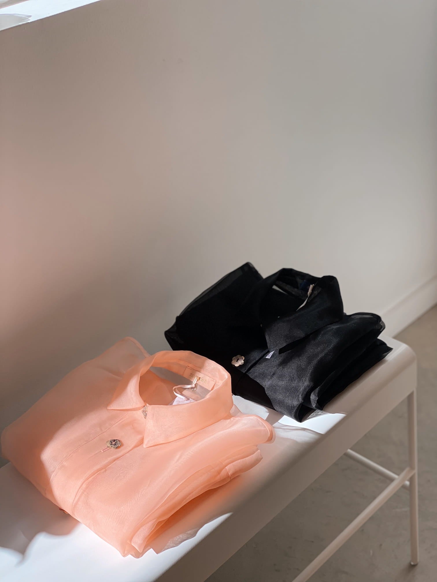 Two folded shirts, including a Sophia Classic Shirt Dress by Noémiah in an oversized fit, are neatly placed on a white bench under soft lighting. One shirt is crafted from luxurious light peach silk organza, while the other is black.