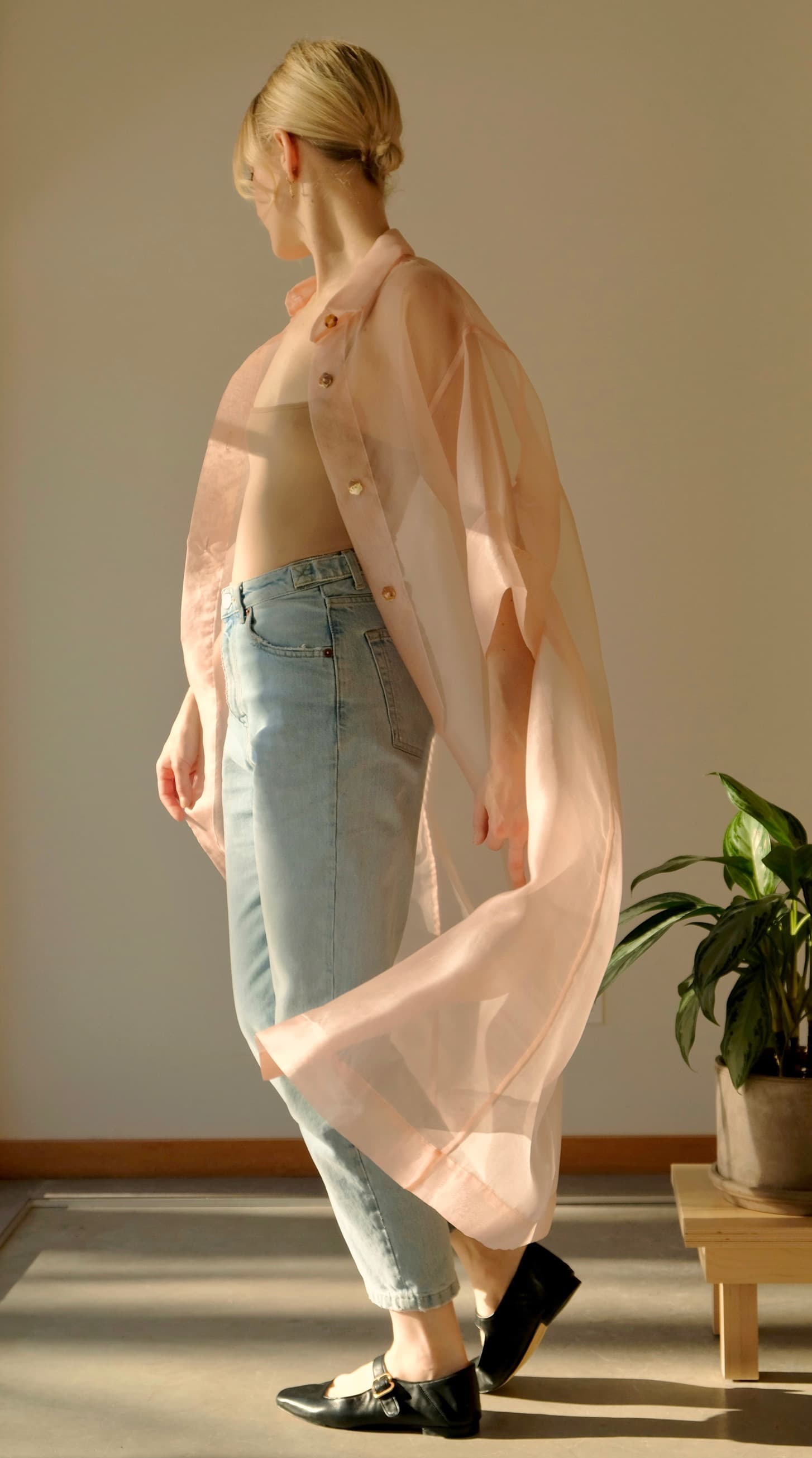 A person stands in a room wearing the sheer Sophia Classic Shirt Dress by Noémiah and light blue jeans. Sunlight casts soft shadows, while a green potted plant on the floor nearby enhances the tranquil scene.