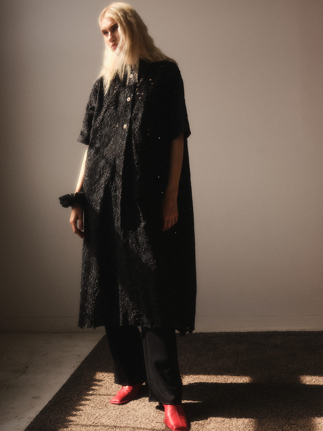 A person with long blonde hair stands in a sunlit room, wearing a textured black cape over black pants and red shoes. The neutral tone is complemented by the oversized Sophia Eyelet Shirt Dress from Noémiah, as light creates a shadow across the floor.