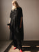 A person with long blonde hair stands in a sunlit room, wearing a textured black cape over black pants and red shoes. The neutral tone is complemented by the oversized Sophia Eyelet Shirt Dress from Noémiah, as light creates a shadow across the floor.