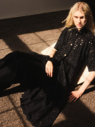 In a sunlit room, a person with long blonde hair sits on the floor wearing the Sophia Eyelet Shirt Dress by Noémiah, its mother of pearl shell buttons adding elegance to the black outfit. The warm light casts shadows, accentuating the texture of the fabric.