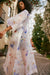 A person confidently stands on a brick pathway wearing the Sophia Printed Shirt Dress by Noémiah, adorned with a sheer floral pattern and mother of pearl buttons. The scene is vibrant and stylish, set against a backdrop of residential buildings and lush greenery.