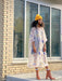 A person with curly hair, wearing a yellow beanie and the Noémiah Sophia Printed Shirt Dress featuring mother of pearl buttons over jeans, stands in front of a brick building with large windows, paired perfectly with brown ankle boots.