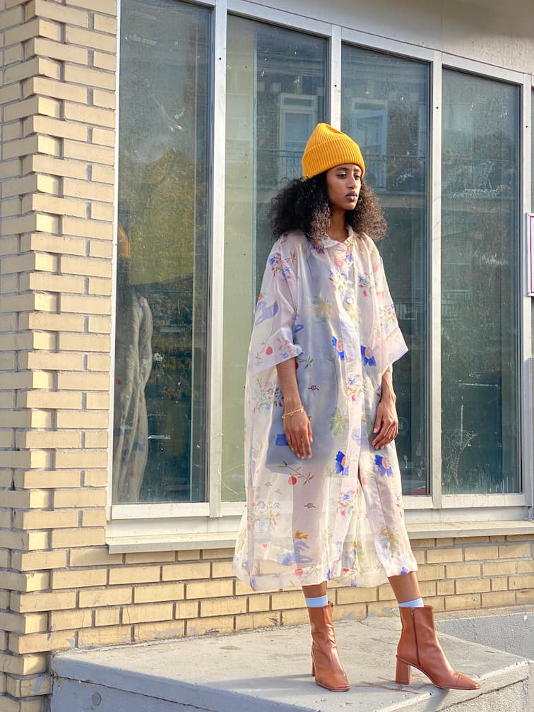 A person with curly hair, wearing a yellow beanie and the Noémiah Sophia Printed Shirt Dress featuring mother of pearl buttons over jeans, stands in front of a brick building with large windows, paired perfectly with brown ankle boots.