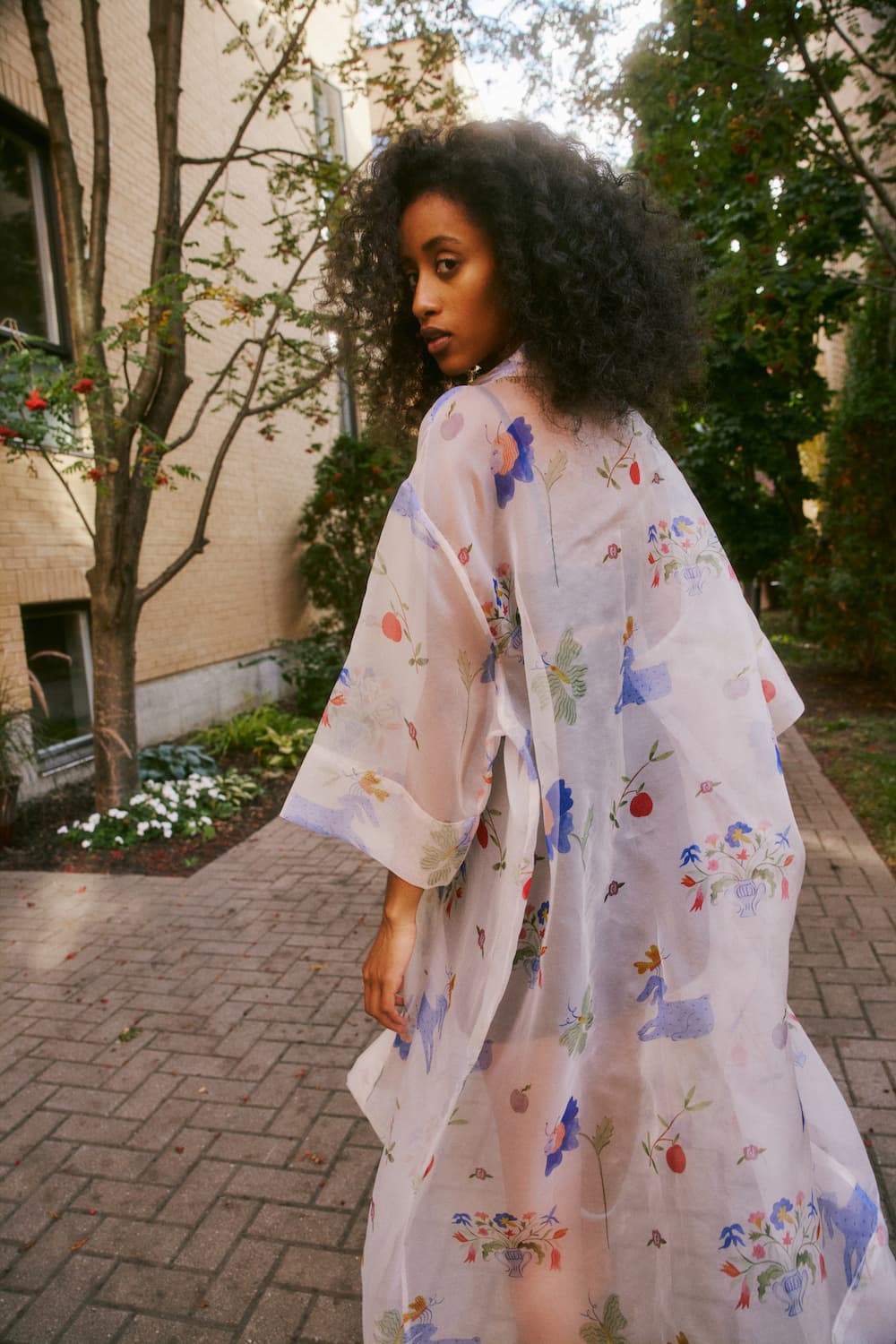 A woman with curly hair is wearing the Sophia Printed Shirt Dress by Noémiah. She stands on a paved path outside, trees and a building gracefully framing the scene.