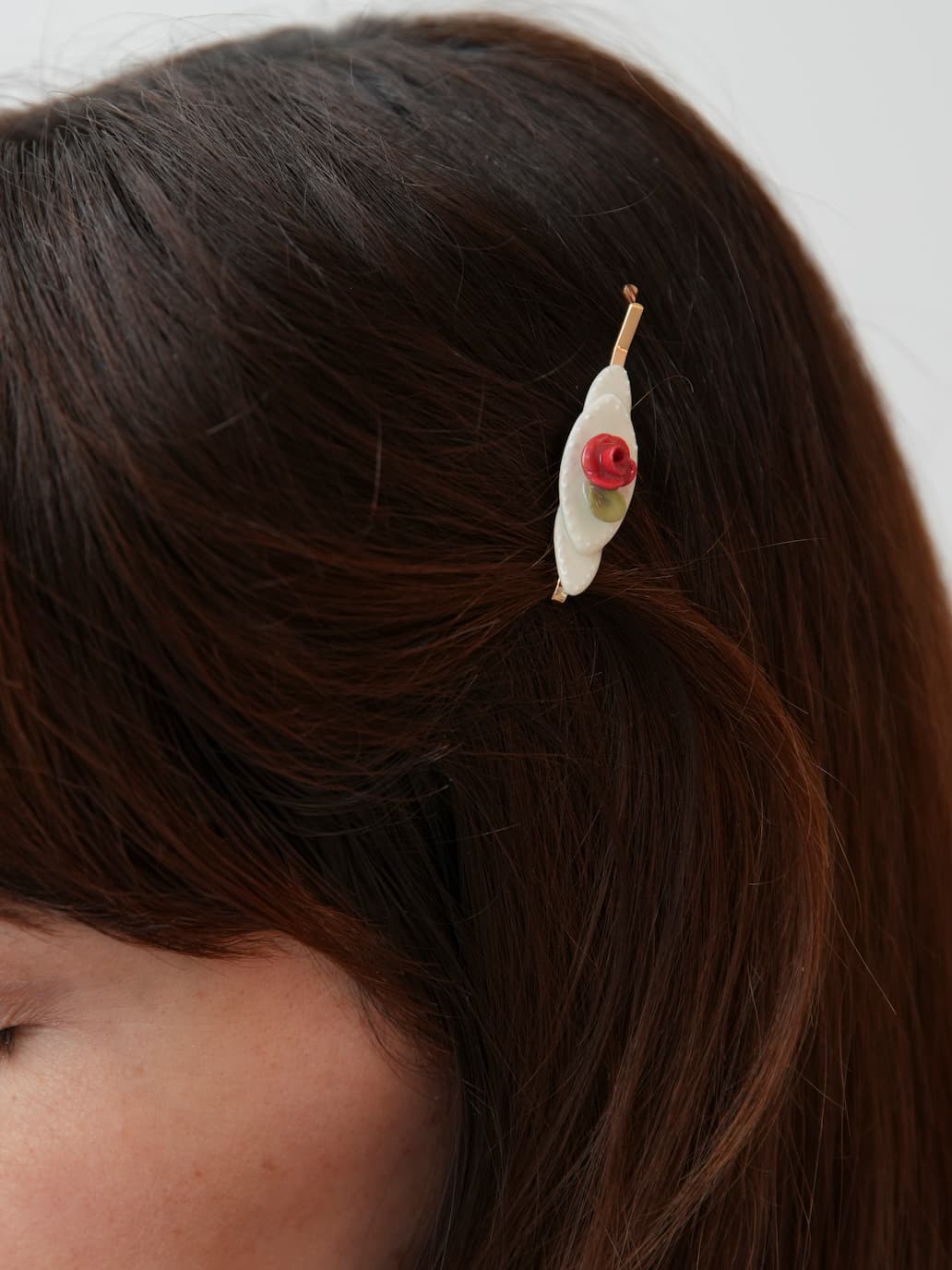 A close-up of brown hair styled with Three Hair Clips by Noémiah, featuring handmade porcelain accents—a white leaf and a red rose. The accessory is in focus, while the person’s face appears partially visible.