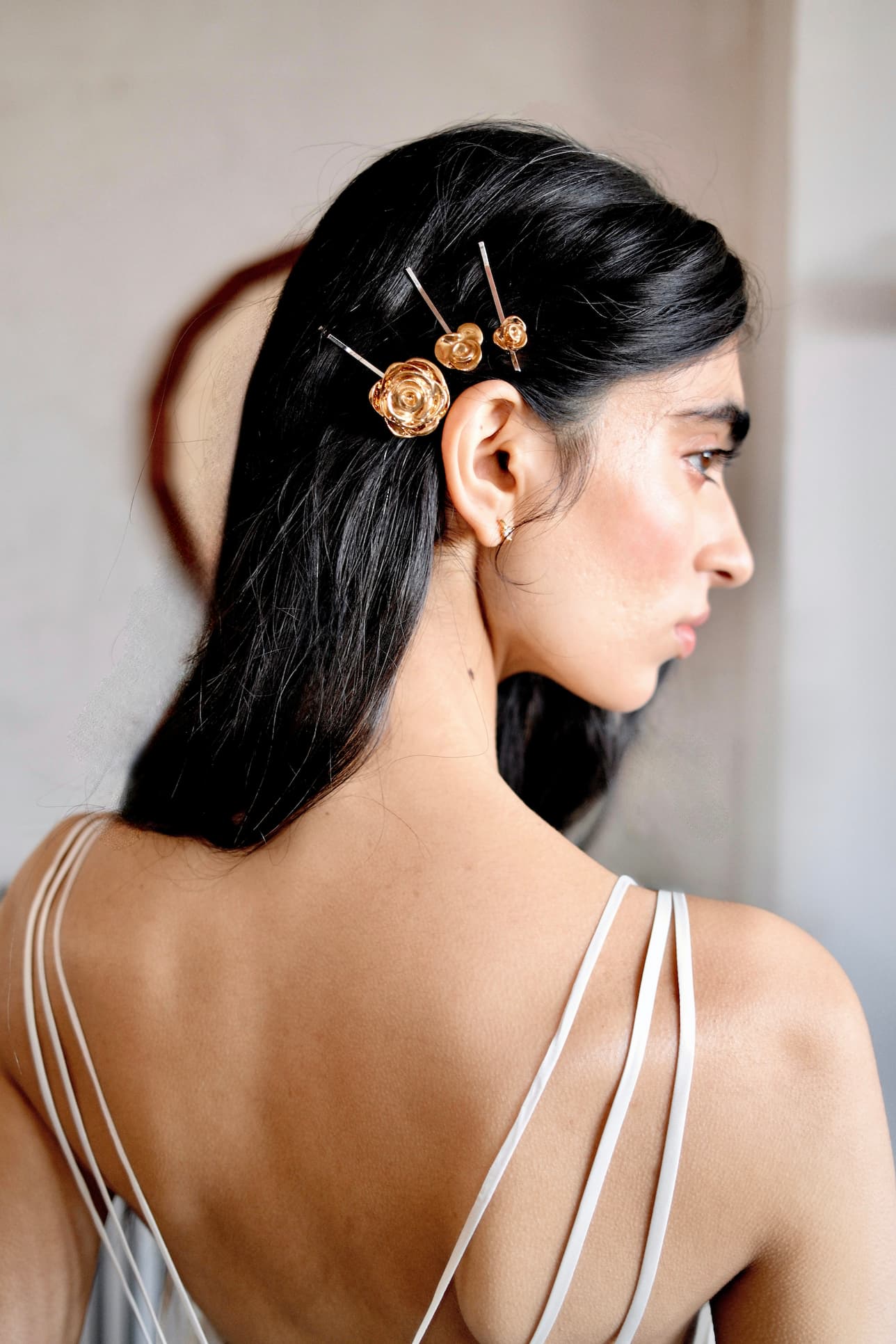 A woman with long dark hair, shown from the back and side in soft lighting, wears Noémiah’s Three Hair Clips—22-karat gold floral designs—on one side, paired with a strappy light-colored dress.