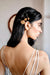 A woman with long dark hair, shown from the back and side in soft lighting, wears Noémiah’s Three Hair Clips—22-karat gold floral designs—on one side, paired with a strappy light-colored dress.