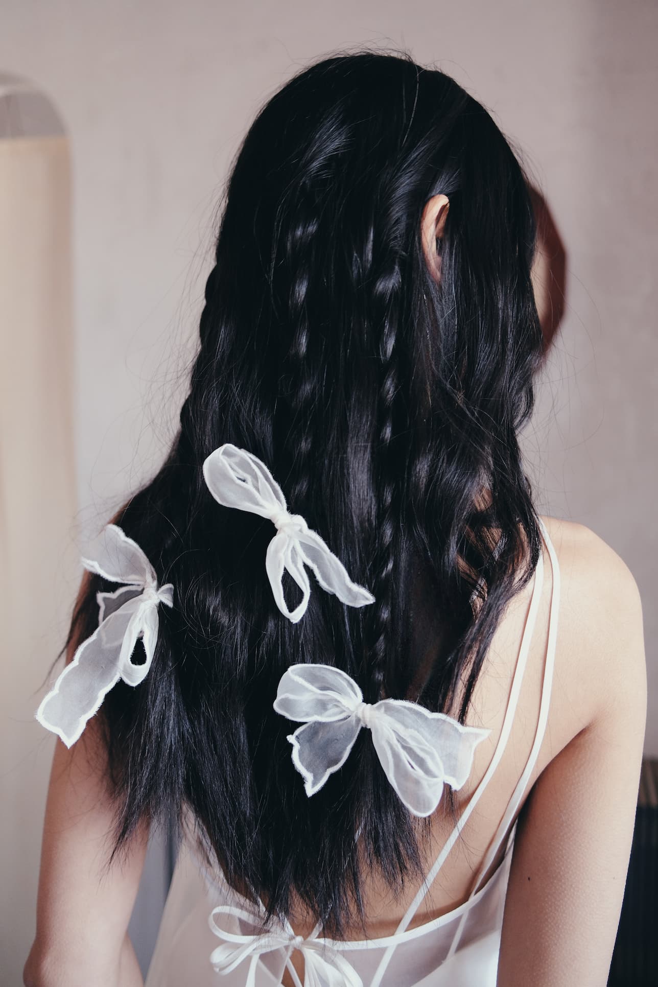 A woman with long, dark, wavy hair stands facing away, wearing a white spaghetti-strap top. Her hair is styled in loose waves and braids adorned with Noémiah’s Three Ribbons—elegant accessories perfect for a wedding hairstyle.