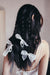 A woman with long, dark, wavy hair stands facing away, wearing a white spaghetti-strap top. Her hair is styled in loose waves and braids adorned with Noémiah’s Three Ribbons—elegant accessories perfect for a wedding hairstyle.