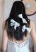 A woman with long, dark braided hair wears Noémiahs Three Ribbons—large scalloped embroidered ribbons—as elegant hair accessories, styled with a sheer white dress and ribbon ties, photographed from behind.