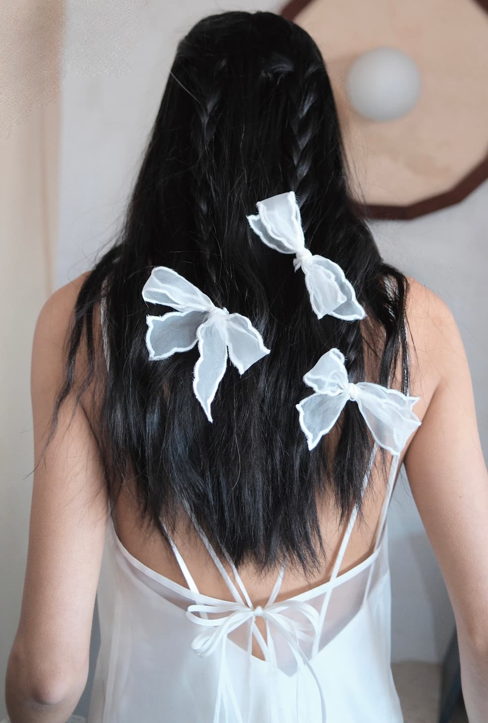 A woman with long, dark braided hair wears Noémiahs Three Ribbons—large scalloped embroidered ribbons—as elegant hair accessories, styled with a sheer white dress and ribbon ties, photographed from behind.
